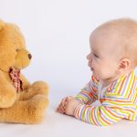 smiling baby girl 6 months old lies on a white isolated background in a bright bodysuit in front of a soft Teddy bear, space for text