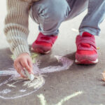 little boy in beige jumper with hood, autumn day in summer in city, draws sun on asphalt with colorful chalks. Creative concept idea and creativity. Background green trees.