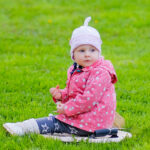 Little baby girl in a pink jacket and hat sits on the grass and looks back