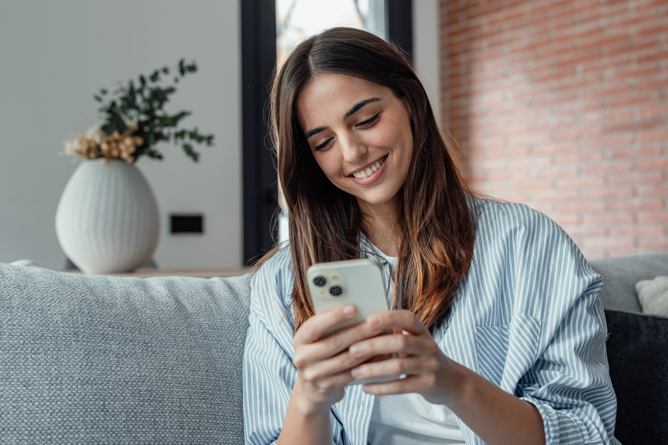 Smiling woman looking at smartphone screen, sitting on couch, enjoying leisure time with mobile device at home, happy young female chatting in social network or surfing internet, shopping online